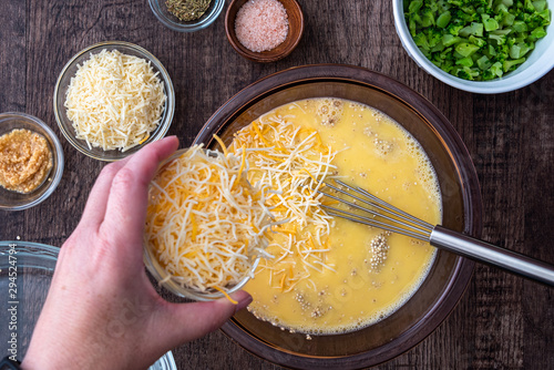 Ingredients for quinoa egg bake, raw egg mix in glass bowl with metal whisk, quinoa, cheese, herbs, broccoli, glass baking dish, on a wood table photo