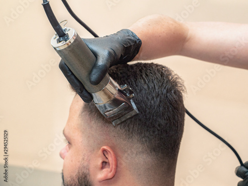 Hairdresser working with clipper, close up view. Hair cutting process. Masters hands in black rubber gloves. Photographed in barbershop. Selective soft focus. Blurred background