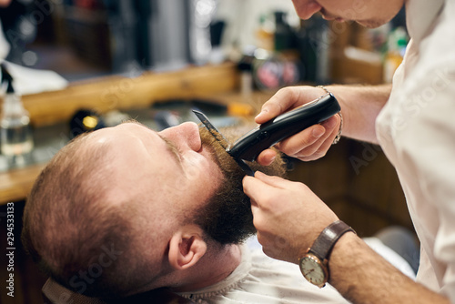 Professional barber using of electric trimmer machine for styling mustaches and beard. Accuracy and concentration of professional barber. Work process in modern hairdressing salon for men.