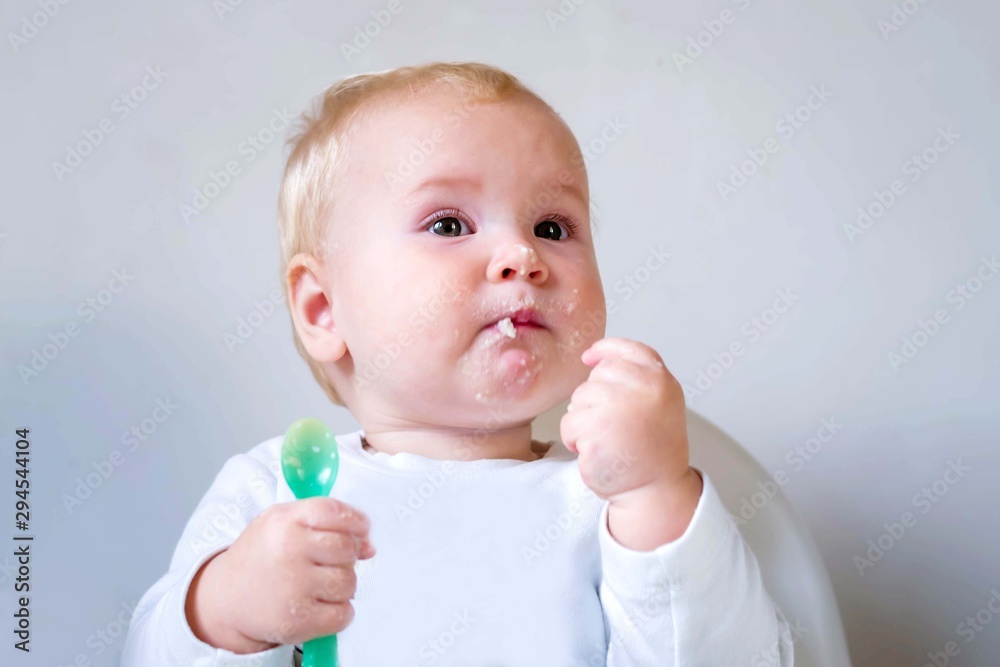Baby, little girl eating lunch with green plastic spoon. Delicious food. Baby eating with pleasure. White plastic chair for feeding.