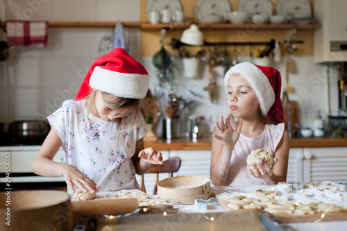 Children are cooking Christmas cookies in cozy home kitchen. Cute kids in santa hats preparation holiday pastry for family. Little girls play with dough. Lifestyle moment. Children chef concept.