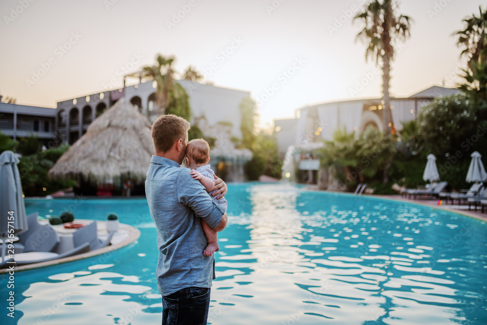 Rear view of happy Caucasian dad hugging his adorable 6 months old son while standing next to swimming pool.