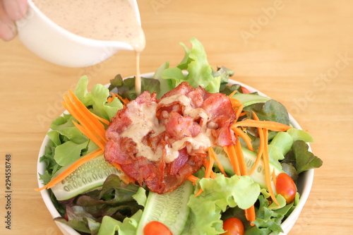 Close up fresh vegetable salad (lettuce, carrot, cucumber, tomato)  with fried bacon and pour by roasted sesame Japanese dressing in white bowl on wooden table background
