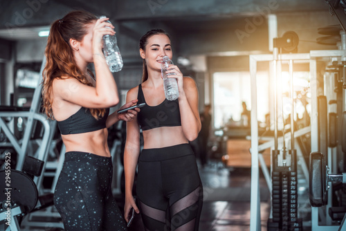Two young attractive Caucasian woman talking together and using smart phone or cellphone while drinking water and relaxation after hard workout at gym