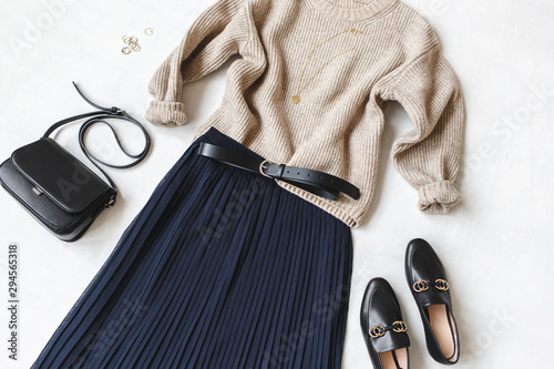 Blue midi pleated skirt, beige knitted sweater, small black cross body bag, belt, loafers (flat shoes) on grey background. Overhead view of women's casual day outfit. Flat lay, top view. Women clothes photo