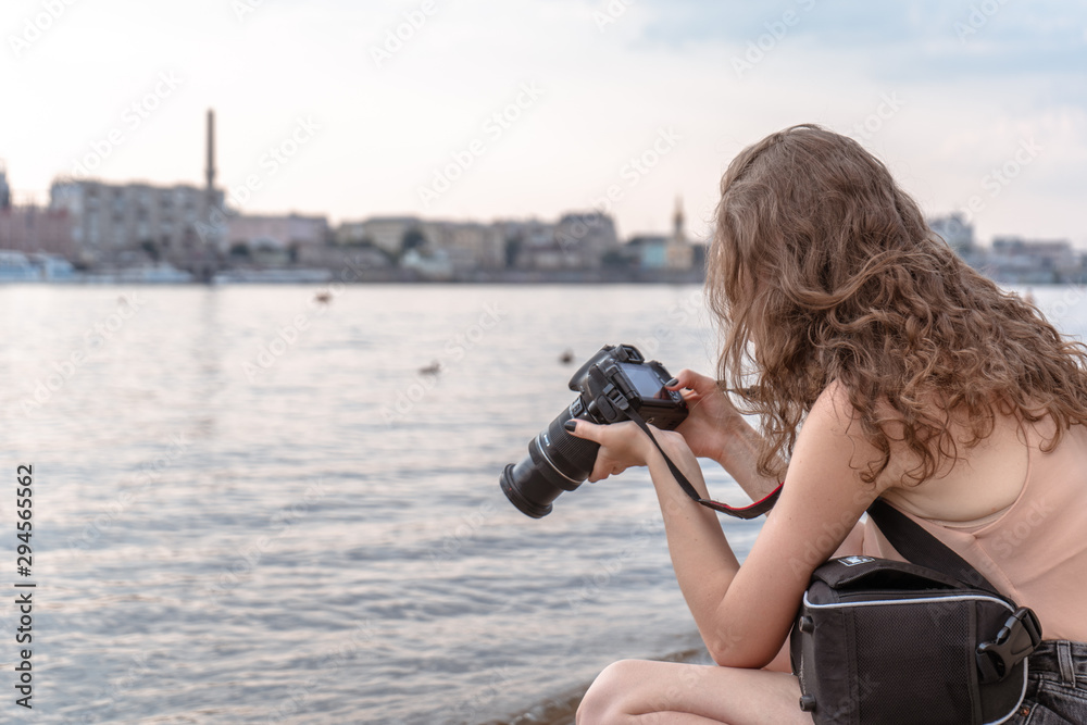 Young woman on the river beach taking photo of the city at sunset with digital camera. Looking at the camera Young woman on the river beach taking photo of the city at sunset with digital camera. Looking at the camera