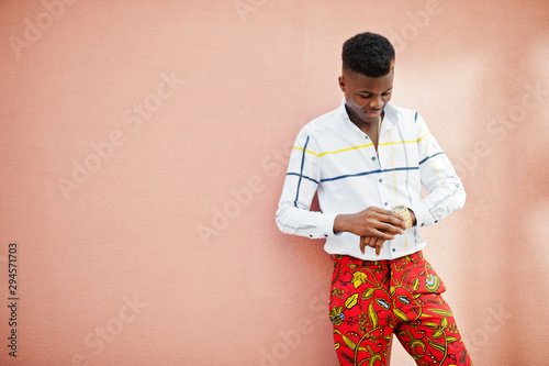 Portrait of handsome stylish african american model man in red throusers and white shirt against pink wall.