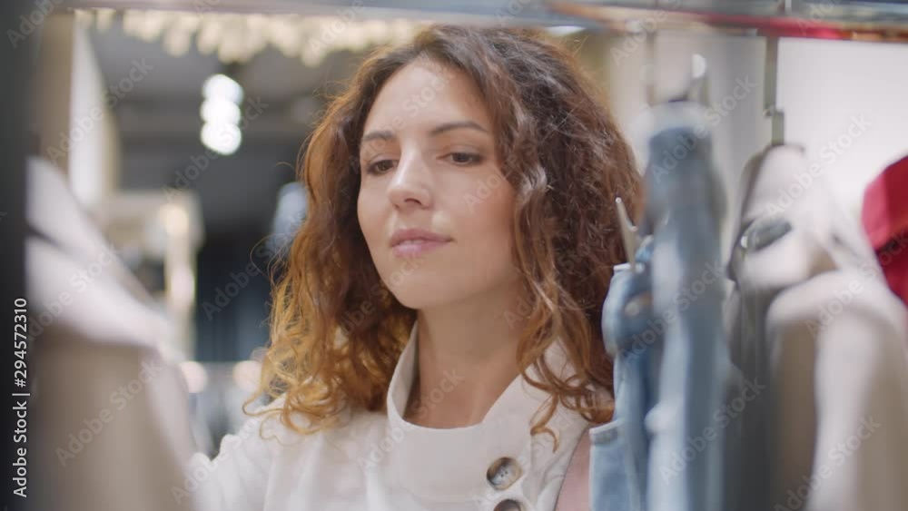 Close up of pretty Caucasian woman with long curly hair standing at rack in store and looking through clothes then taking hanger with jacket and attaching it to herself