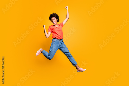 Full size photo of crazy funky delighted lady jump celebrate victory in lottery raise her fists scream yeah wear red t-shirt denim jeans sneakers isolated over yellow color background