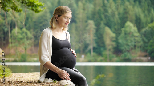 Beautiful, cute pregnant woman sits near a mountain lake in a forest on the shore