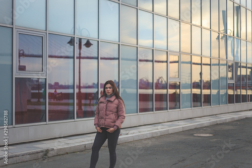 Young tired pretty pregnant girl in pink warm down jacket. Some reflection in the window. Late autumn, the cold is coming.
