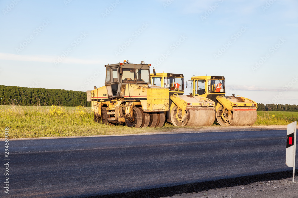 Three large yellow asphalt compactor standing on the side of the track during highway road repair and laying black new asphalt against a landscape with a blue sky and clouds in the summer.
