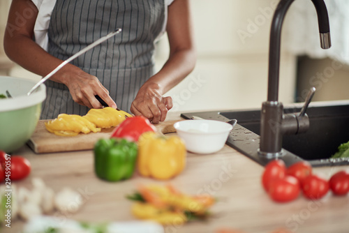 Close up of mixed race woman in apron cutting yellow pepper for dinner. Kitchen interior. On kitchen counter are peppers, zucchini and tomatoes.