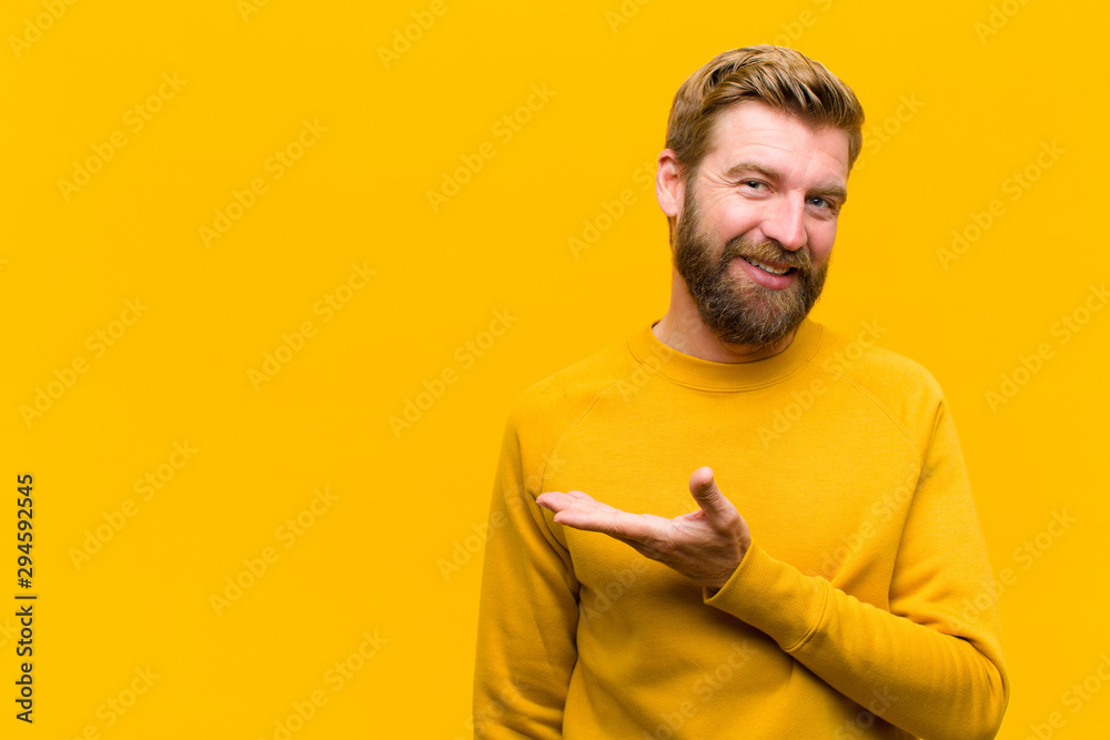 young blonde man smiling cheerfully, feeling happy and showing a concept in copy space with palm of hand against orange wall - obrazy, fototapety, plakaty 