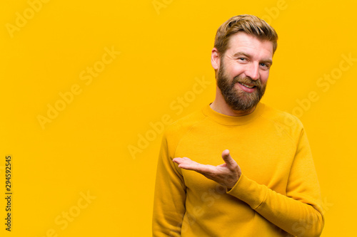 young blonde man smiling cheerfully, feeling happy and showing a concept in copy space with palm of hand against orange wall