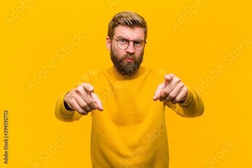 young blonde man pointing forward at camera with both fingers and angry expression, telling you to do your duty against orange wall