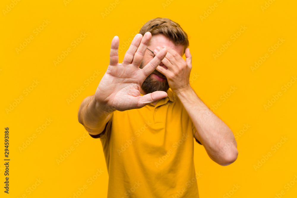 young blonde man covering face with hand and putting other hand up front to stop camera, refusing photos or pictures against orange wall - obrazy, fototapety, plakaty 