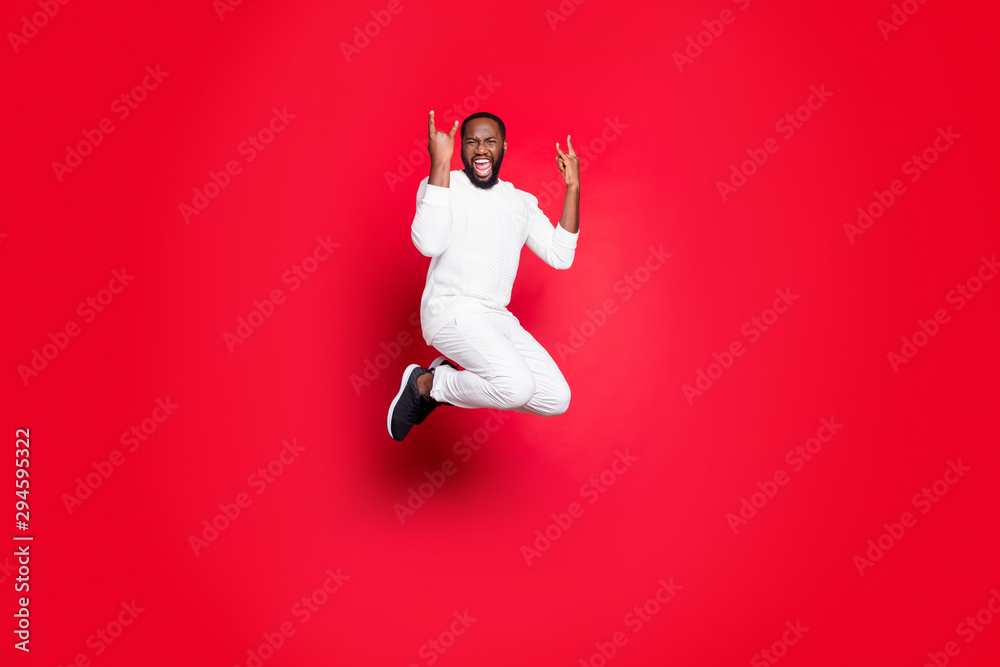 Full size photo of excited dark skin handsome guy jumping high rejoicing at rock concert showing horns hands wear white sweater trousers isolated red background Full size photo of excited dark skin handsome guy jumping high rejoicing at rock concert showing horns hands wear white sweater trousers isolated red background