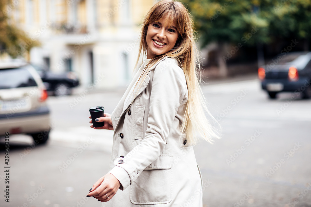 The concept of street fashion. portrait of Young girl dressed in beige trench coat. Posing against the window of the boutique girl smiling and drinking coffee