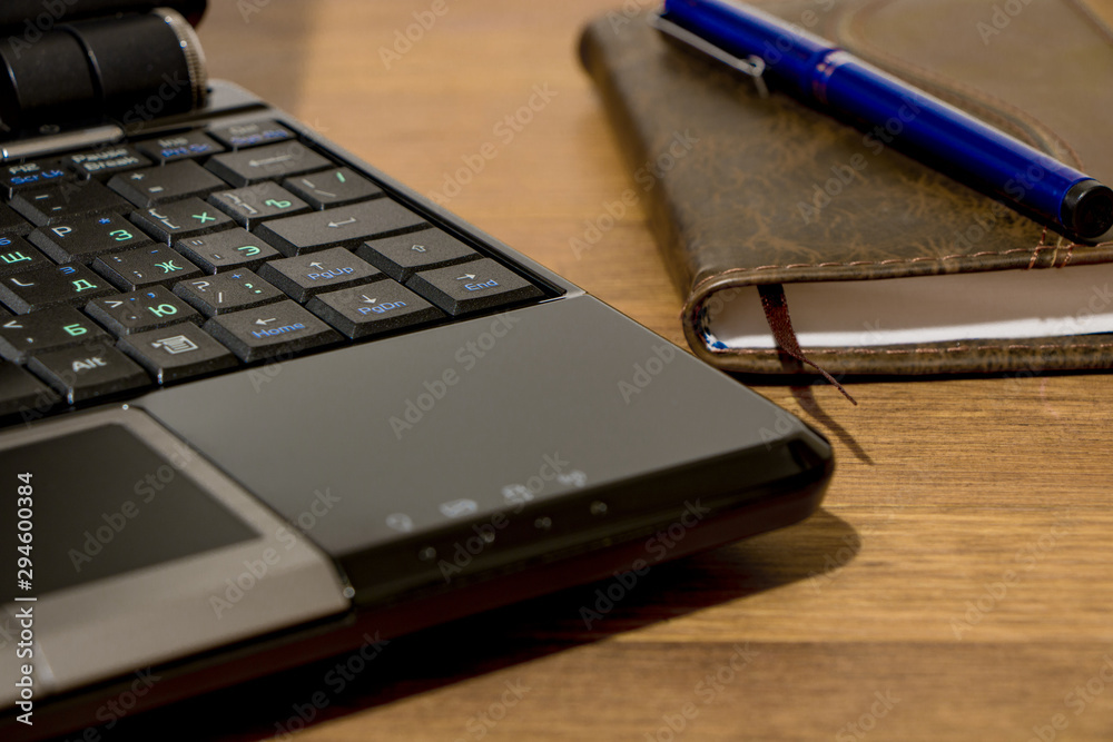 Remote workplace. Remote work environment. View of a natural wood table with several items, a black laptop, a diary and a pen. Remote workplace. Remote work environment. View of a natural wood table with several items, a black laptop, a diary and a pen.