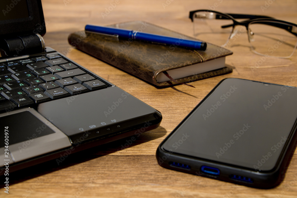 Remote workplace. Remote working environment. View of a natural wood table with several items, a black laptop, a diary, a pen, glasses and a mobile phone. Remote workplace. Remote working environment. View of a natural wood table with several items, a black laptop, a diary, a pen, glasses and a mobile phone.