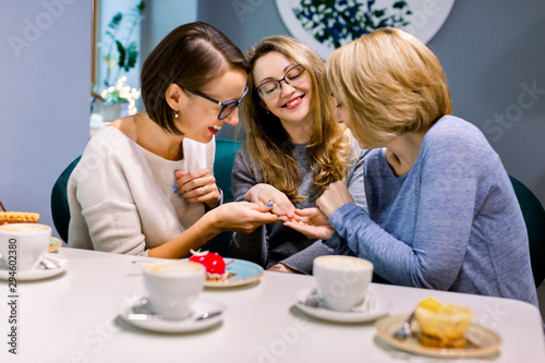 Looking at ring. Blonde-haired and dark-haired women holding hand of their pretty friend looking at engagement ring. Women in cafe indoors