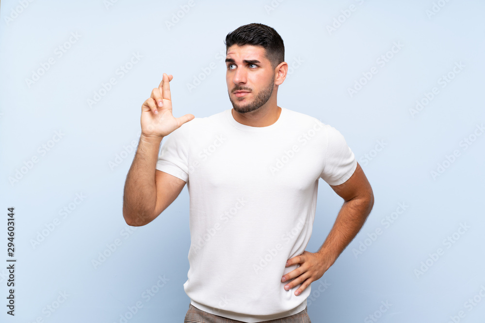 Fototapeta premium Caucasian handsome man over isolated blue background with fingers crossing and wishing the best