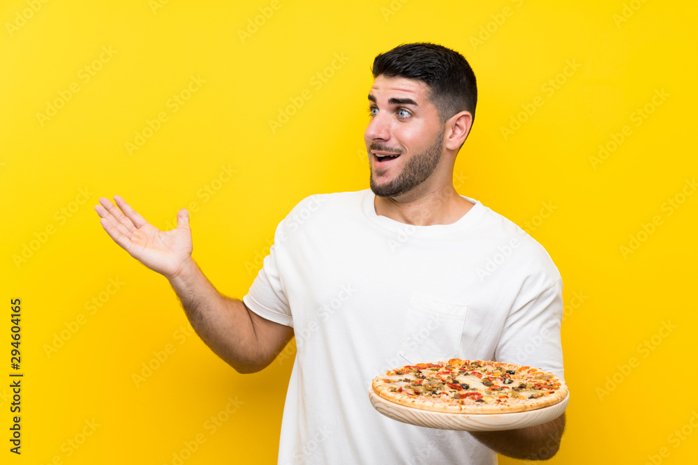 Young handsome man holding a pizza over isolated yellow wall with surprise facial expression