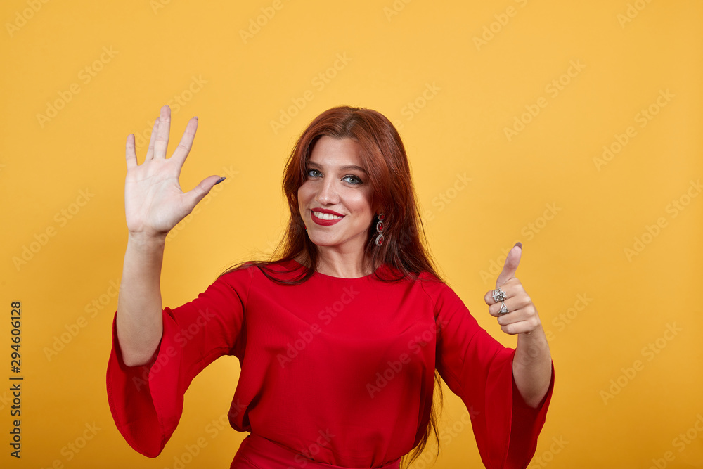 Obraz premium Cheerful girl showing gestures by both hands. Her left thumb is up. Woman holding right hand up. Wearing red dress smiling person stands on colourful background. Brunette with red lipstick looks happy