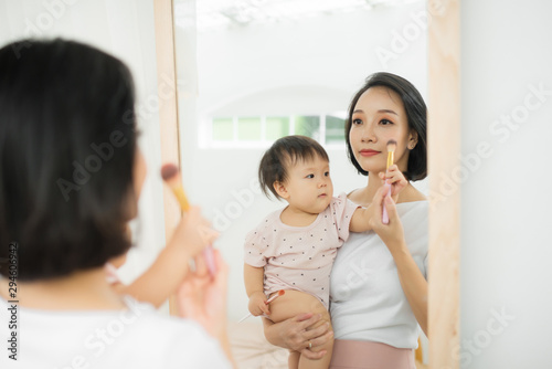 Funny family at home. Mother and her child girl are doing your makeup and having fun near mirror. Baby girl explores mother's cosmetics at home