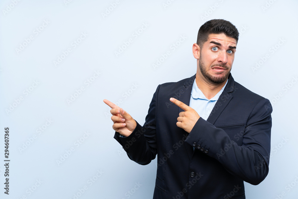 Handsome man over isolated blue background frightened and pointing to the side Handsome man over isolated blue background frightened and pointing to the side