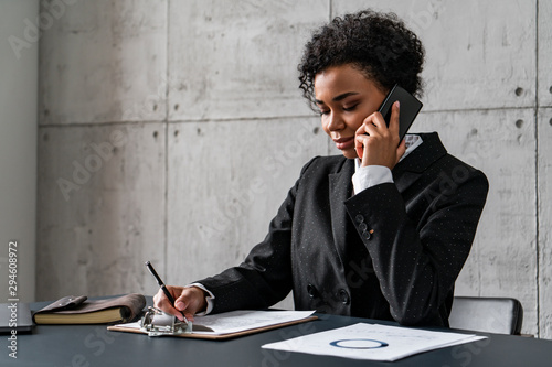 Side view of young African American businesswoman talking on smartphone and taking notes in blurred loft office. Concept of business communication photo