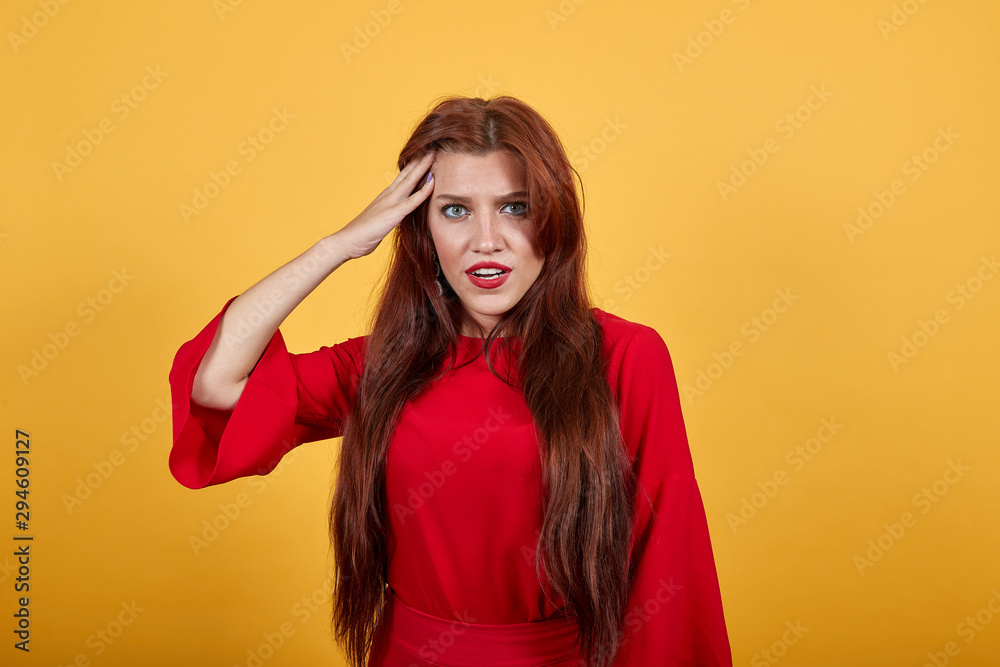 Pretty woman holding her right hand near temple shows by that gesture confusion. Puzzled girl in red garment with long, dark, shining hair stands on orange background.