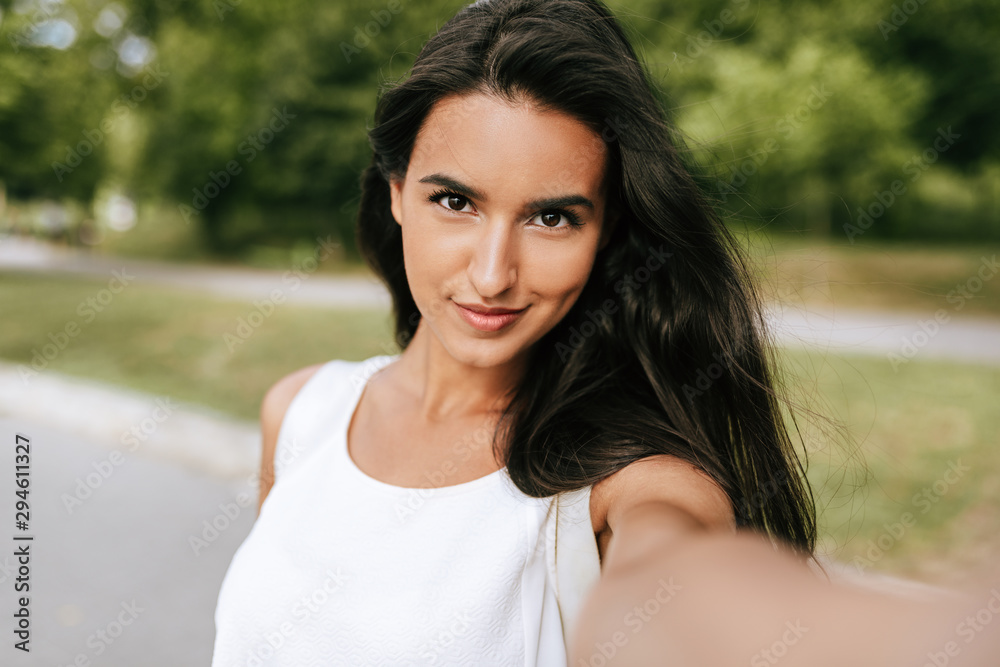Close-up portrait of a beautiful young brunette smiling woman taking self portrait on nature background in the park. Pretty female making selfie on her device outdoors. People, lifesetyle and travel