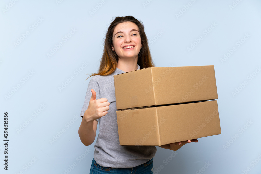 Young brunette girl over isolated blue background holding a box to move it to another site with thumb up Young brunette girl over isolated blue background holding a box to move it to another site with thumb up