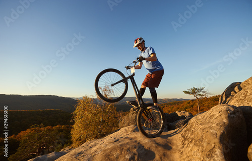 Fearless cyclist balancing on back wheel on trial bike. Sportsman rider making acrobatic stunt on the edge of big boulder on the top of mountain at sunset. Concept of extreme sport active lifestyle
