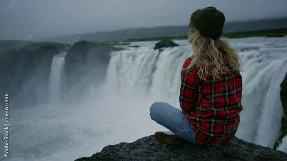 Calm and tranquil young woman enjoys view over epic waterfall during midnight sun in norway or iceland. Scandinavian adventure concept, exploring natural wonders in great outdoors