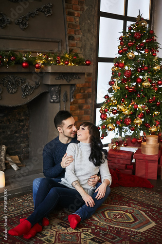 The young happy couple, in warm sweaters and red socks, hugging and sitting on the authentic carpet near the fireplace and Christmas tree. Christmas celebration and New year holidays © innarevyako