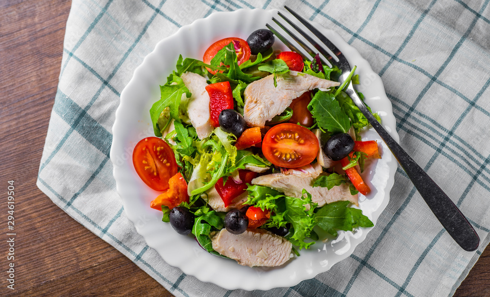 Fresh salad with chicken breast, arugula, black olives,red pepper, lettuce, fresh sald leaves and tomato on a white plate on wooden table background