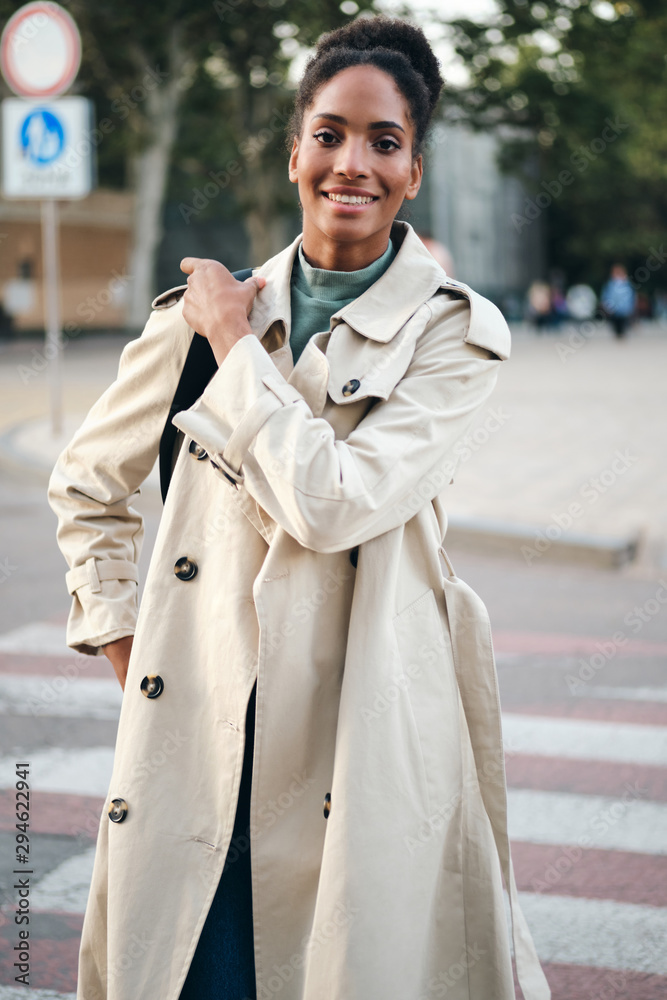 Beautiful smiling casual African American girl in stylish trench coat  joyfully looking in camera while walking down the street Stock-Foto | Adobe  Stock