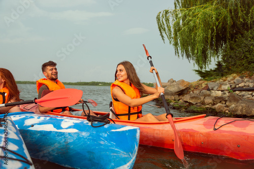 Happy young caucasian group of friends kayaking on river with sunset in the backgrounds. Having fun in leisure activity. Happy male and female model laughting on the kayak. Sport, relations concept. © master1305