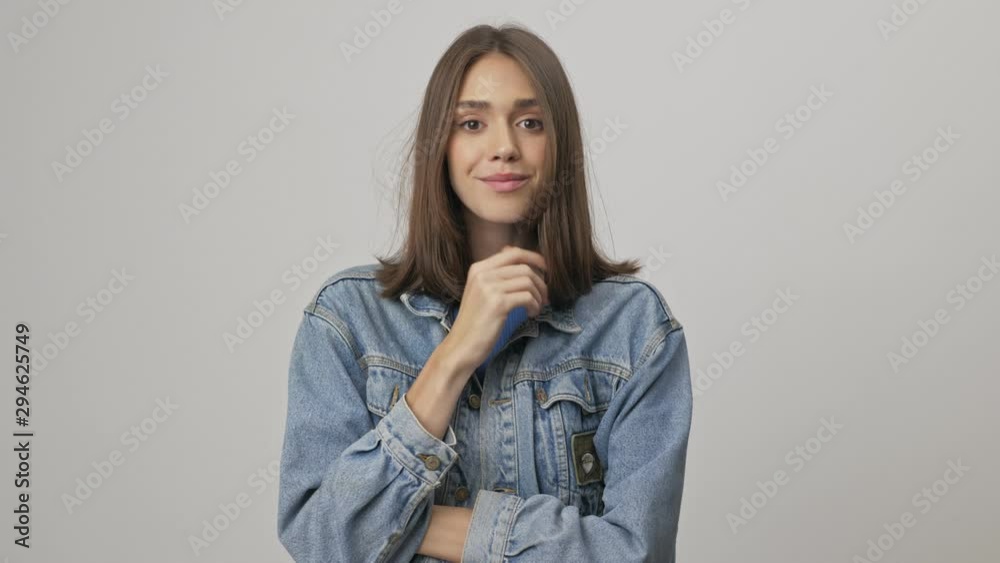 Cute young brunette woman in denim jacket smiling and touching her chin while looking at the camera over gray background isolated Cute young brunette woman in denim jacket smiling and touching her chin while looking at the camera over gray background isolated