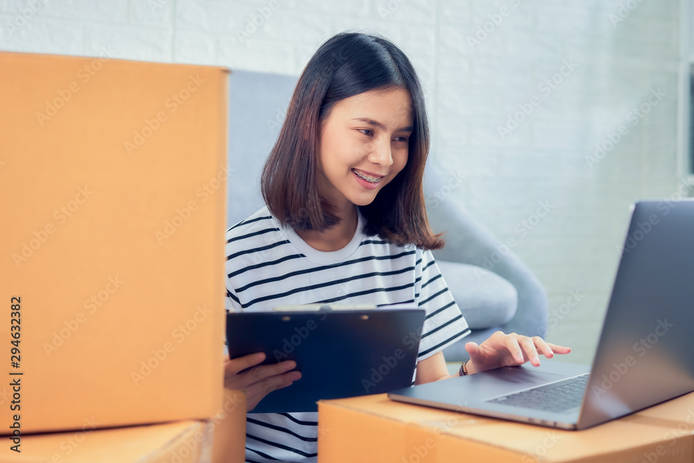 Smiling Asian woman owner check the customer order from the computer, seller prepares the delivery box. Startup small business concept. Smiling Asian woman owner check the customer order from the computer, seller prepares the delivery box. Startup small business concept.