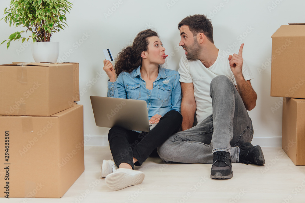 Woman wants to use a credit card to buy something online. Her husband is arguing. Sheis showing him tongue. Young couple with laptop in the apartment, sitting on the floor, surfing in the internet. Woman wants to use a credit card to buy something online. Her husband is arguing. Sheis showing him tongue. Young couple with laptop in the apartment, sitting on the floor, surfing in the internet.