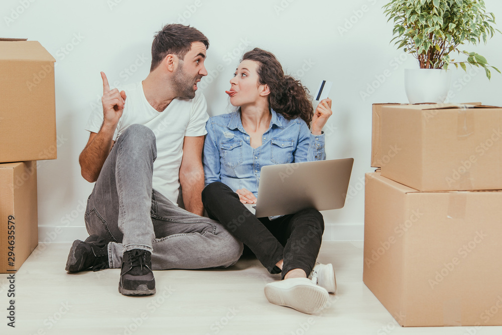 Woman wants to use a credit card to buy something online. Her husband is arguing. Sheis showing him tongue. Young couple with laptop in the apartment, sitting on the floor, surfing in the internet. Woman wants to use a credit card to buy something online. Her husband is arguing. Sheis showing him tongue. Young couple with laptop in the apartment, sitting on the floor, surfing in the internet.