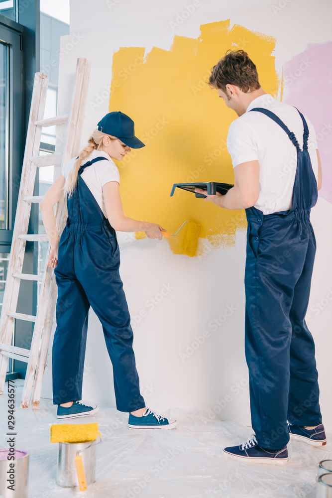 young painter holding roller tray while attractive colleague painting wall with paint roller