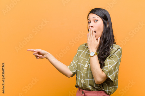 young pretty hispanic woman feeling happy, shocked and surprised, covering mouth with hand and pointing to lateral copy space against brown wall photo