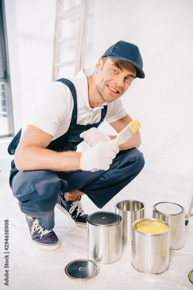 handsome young painter in uniform holding paintbrush near cans with paint and smiling at camera handsome young painter in uniform holding paintbrush near cans with paint and smiling at camera