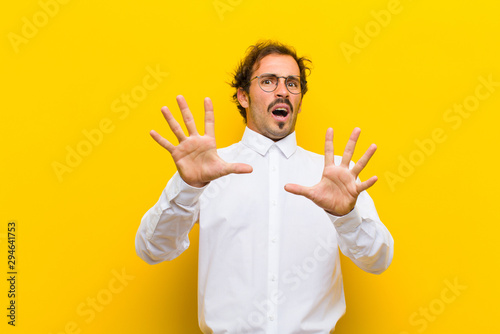 young handsome man feeling terrified, backing off and screaming in horror and panic, reacting to a nightmare against orange wall