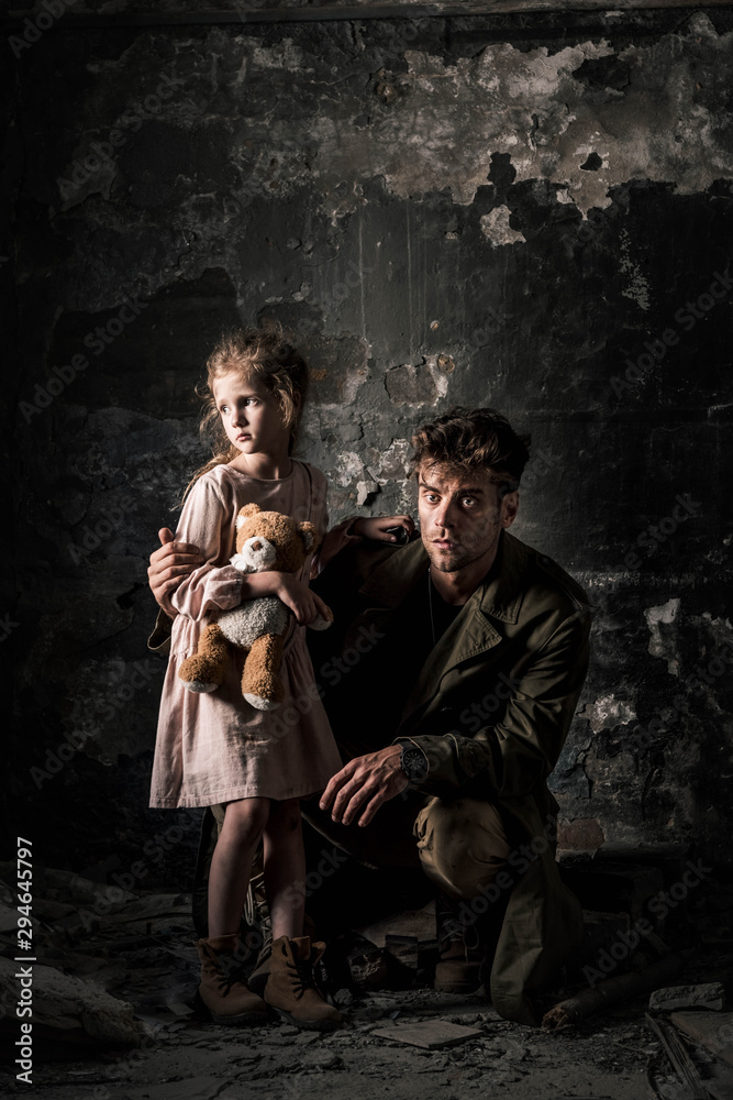 handsome man sitting near upset kid with teddy bear in dirty room, post apocalyptic concept